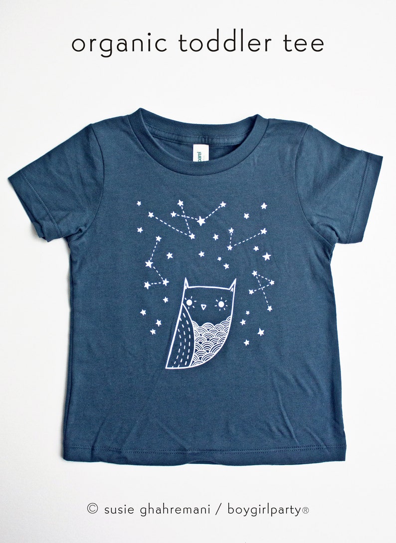 May include: A dark blue organic toddler tee with a white owl and constellation design. The text "organic toddler tee" is printed at the top of the image. The text "susie ghahremani / boygirlparty&reg;" is printed at the bottom of the image.