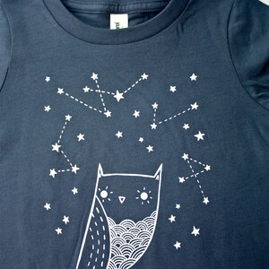 May include: A dark blue t-shirt with a white owl and constellation design. The owl has a whimsical design with large eyes and a wavy pattern on its chest. The constellation is made up of white stars connected by dotted lines.