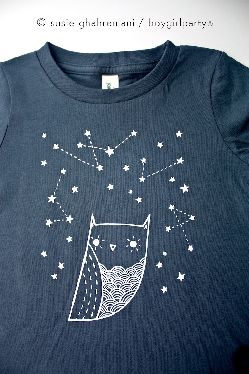 May include: A dark blue t-shirt with a white owl and constellation design. The owl has a whimsical design with large eyes and a wavy pattern on its chest. The constellation is made up of white stars connected by dotted lines.