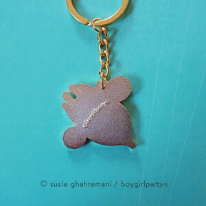 May include: Gold-colored metal keychain featuring a cartoon character with a crown. The back of the keychain has the text "&copy; susie ghahremani / boygirlparty&reg;".