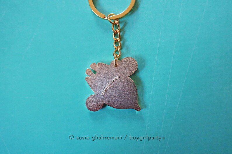 May include: Gold-colored metal keychain featuring a cartoon character with a crown. The back of the keychain has the text "&copy; susie ghahremani / boygirlparty&reg;".