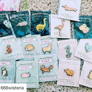 May include: A collection of enamel pins featuring various animal designs, including a fox, a hedgehog, an alpaca, a squirrel, a pig, a seahorse, a platypus, a dinosaur, a flamingo, a pair of birds, a capybara, and a chicken. The pins are packaged on blue and white cards with the text "Boygirlparty.com" and "The artwork of Suzi Ghahremani/Boygirlparty".