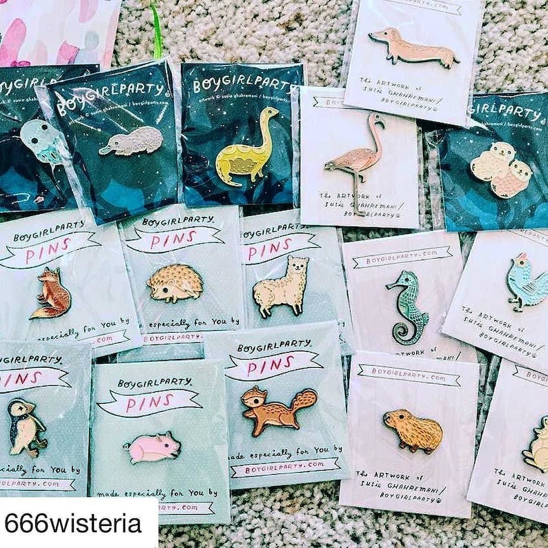 May include: A collection of enamel pins featuring various animal designs, including a fox, a hedgehog, an alpaca, a squirrel, a pig, a seahorse, a platypus, a dinosaur, a flamingo, a pair of birds, a capybara, and a chicken. The pins are packaged on blue and white cards with the text "Boygirlparty.com" and "The artwork of Suzi Ghahremani/Boygirlparty".