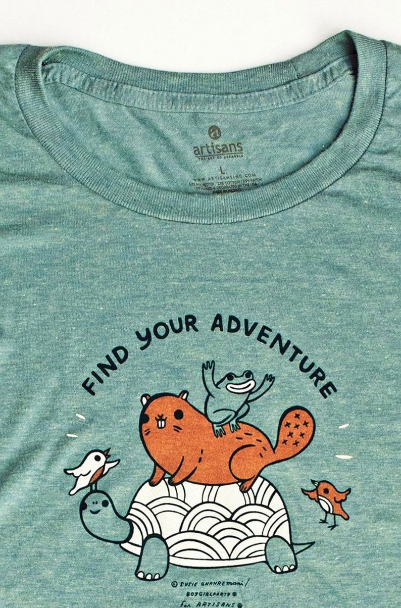 May include: A teal blue t-shirt with a graphic of a turtle, a beaver, a frog, and two birds. The text "Find Your Adventure" is written in a black, curved font around the graphic. The text "Susie Ghanretmon/ BoyGirlParty for Artisans" is printed in black at the bottom of the graphic.
