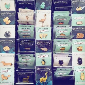 May include: A collection of enamel pins featuring various designs, including animals, plants, and abstract shapes. The pins are displayed on a white background with the text "Boygirlparty" and "Pins" on several of the cards.