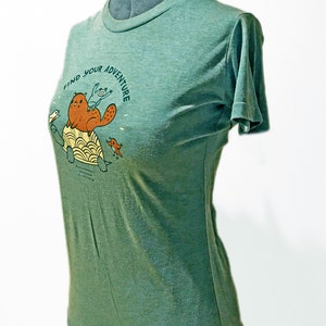 May include: Green short-sleeved t-shirt with a graphic of a beaver riding a turtle with two birds. The text "Find Your Adventure" is printed on the shirt.