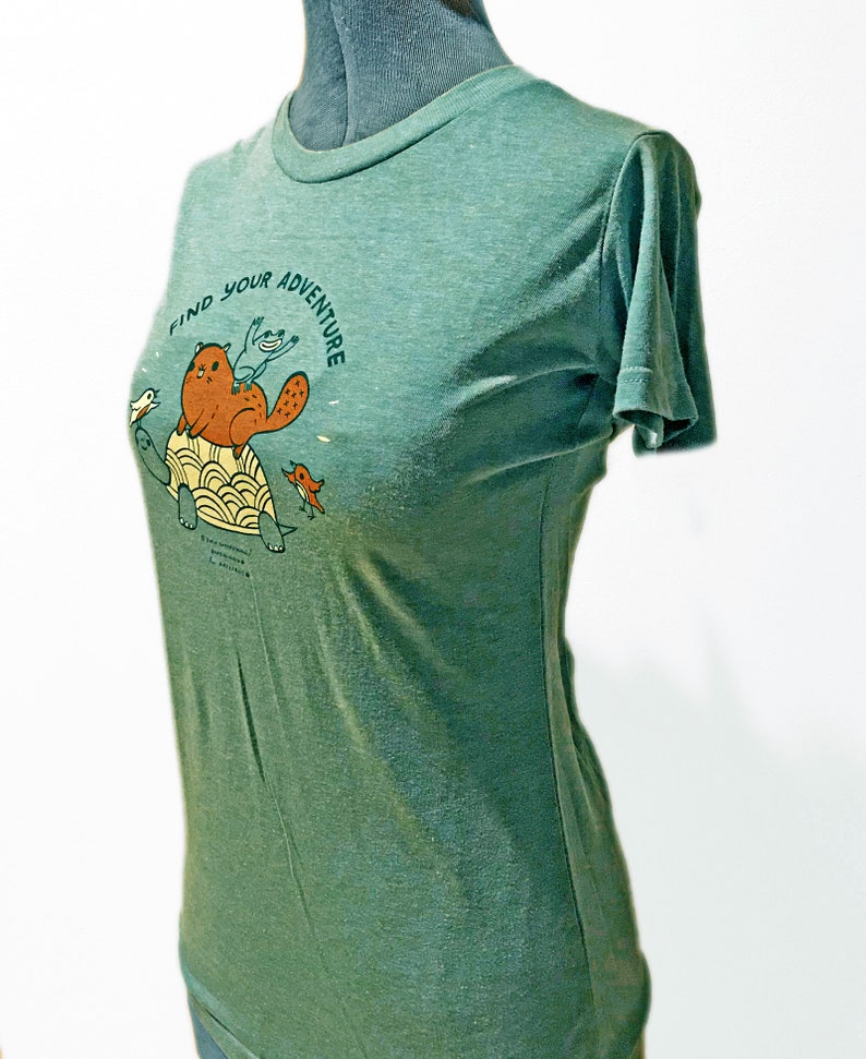 May include: Green short-sleeved t-shirt with a graphic of a beaver riding a turtle with two birds. The text "Find Your Adventure" is printed on the shirt.