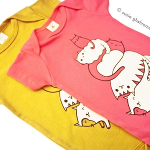 May include: Two baby onesies, one yellow and one pink. The pink onesie has a graphic of cats playing. The yellow onesie has a graphic of cats playing.
