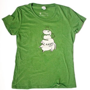 K&ouml;nnte beinhalten: Gr&uuml;nes, umweltfreundliches T-Shirt mit einem Aufdruck von f&uuml;nf &uuml;bereinander gestapelten wei&szlig;en Katzen mit gr&uuml;nen Akzenten. Der Text "susie ghahremani / boygirlparty&reg;" befindet sich am unteren Rand des Shirts.