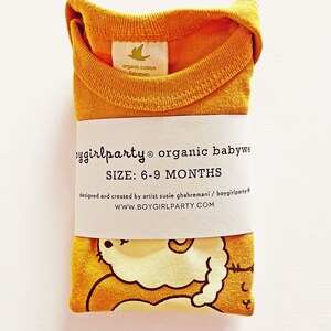 May include: A mustard yellow organic cotton baby onesie with a cute cartoon illustration of a sleeping cat. The onesie is folded and has a label that says "boygirlparty organic babywear" and "SIZE: 6-9 MONTHS".