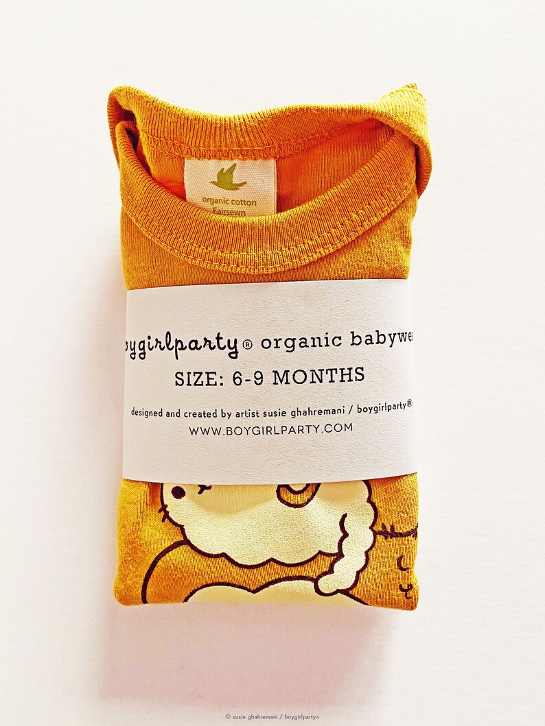 May include: A mustard yellow organic cotton baby onesie with a cute cartoon illustration of a sleeping cat. The onesie is folded and has a label that says "boygirlparty organic babywear" and "SIZE: 6-9 MONTHS".