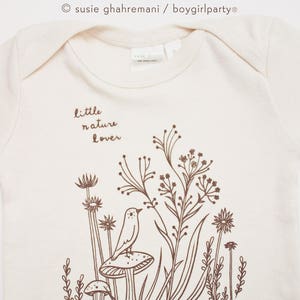 Puede incluir: Un body blanco para beb&eacute; con un gr&aacute;fico marr&oacute;n de un p&aacute;jaro posado en un hongo en un campo de flores silvestres. El texto "little nature lover" est&aacute; impreso en el body.