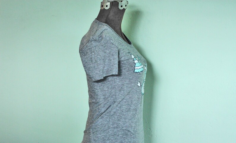 May include: A heather grey t-shirt with a graphic of a cartoon snowman wearing a blue hat and scarf. The snowman is surrounded by green Christmas trees.