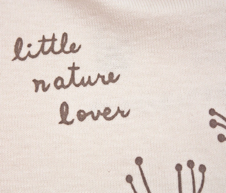 May include: White fabric with brown text that reads "little nature lover". The fabric has a subtle texture and is decorated with small brown floral designs.
