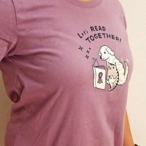May include: A purple t-shirt featuring a graphic of a dog and cat reading a book together. The text on the shirt reads "Let's Read Together!"