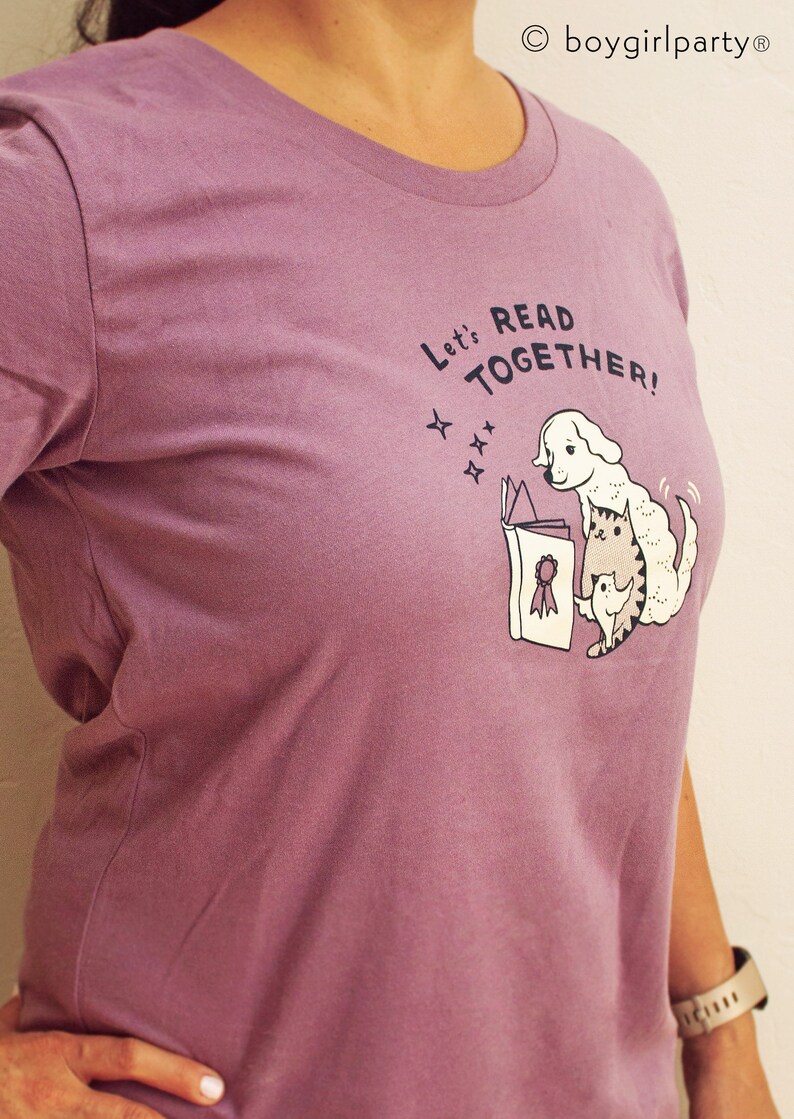 May include: A purple t-shirt featuring a graphic of a dog and cat reading a book together. The text on the shirt reads "Let's Read Together!"