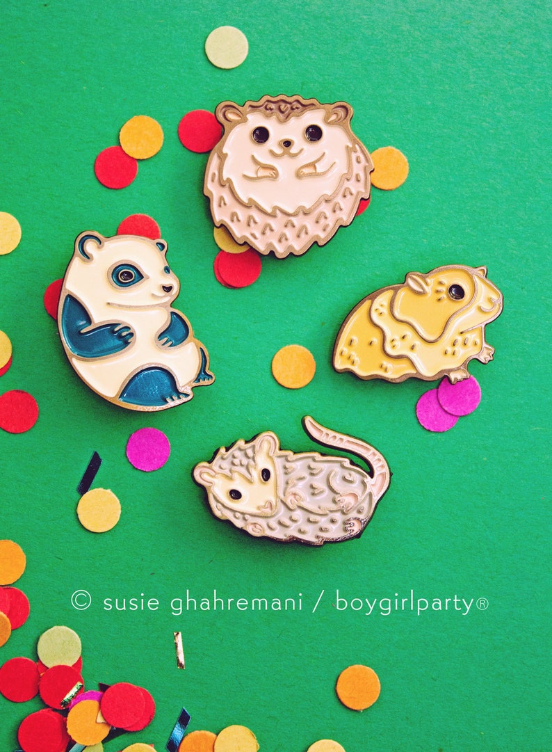 May include: Four enamel pins featuring different animals: a hedgehog, a panda, a hamster, and a possum. The pins are on a green background with colorful confetti.