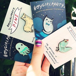 Op de afbeelding: Vier emaillepins op kaarten met de tekst "Boygirlparty" en de naam van de kunstenaar. De pins tonen een lama, een kat, een kwal en een vogel.