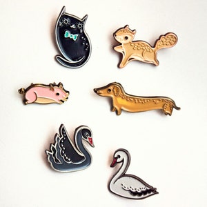 May include: Six enamel pins featuring various animals: a black cat with a bow tie, a brown squirrel, a pink pig, a brown dachshund, a black swan, and a white swan. The pins are arranged on a white background with the text "new pins" at the top.