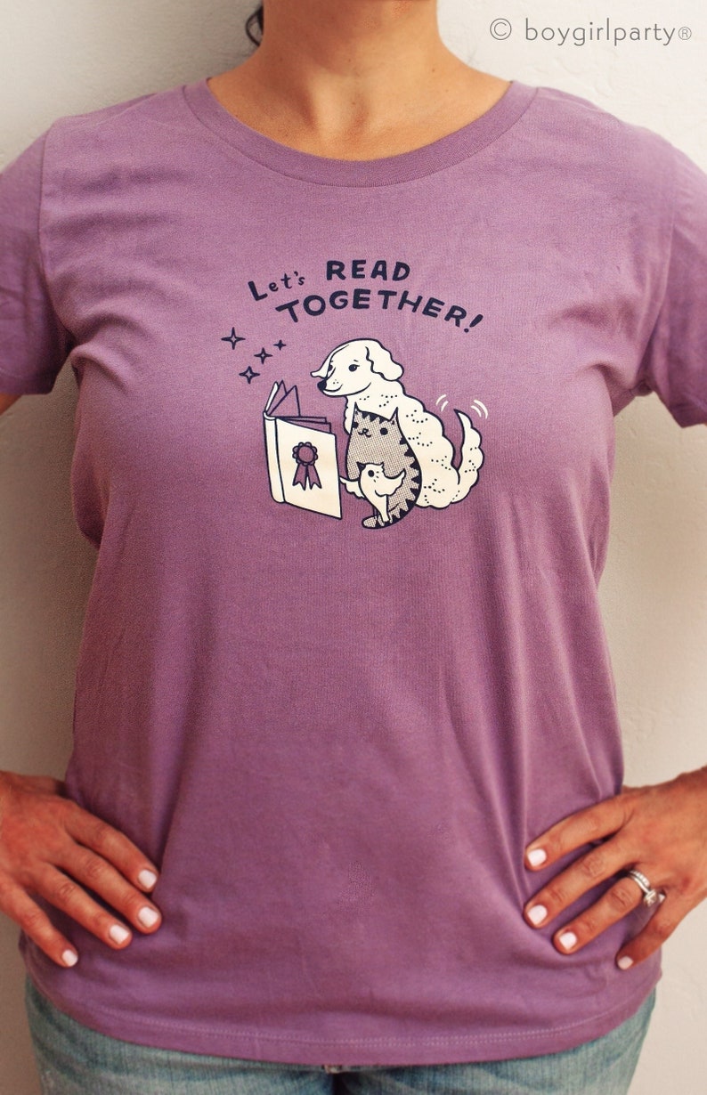 May include: Purple t-shirt featuring a white dog and cat reading a book. The text on the shirt reads "Let's Read Together!"