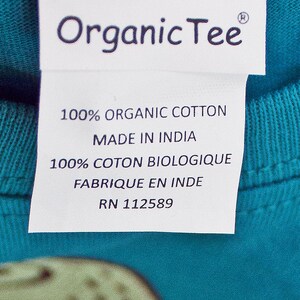 Może przedstawiać: Biała metka z czarnym tekstem na turkusowej koszulce. Metka zawiera napis "Organic Tee", "100% Organic Cotton", "Made in India", "100% Coton Biologique", "Fabrique en Inde" i "RN 112589".