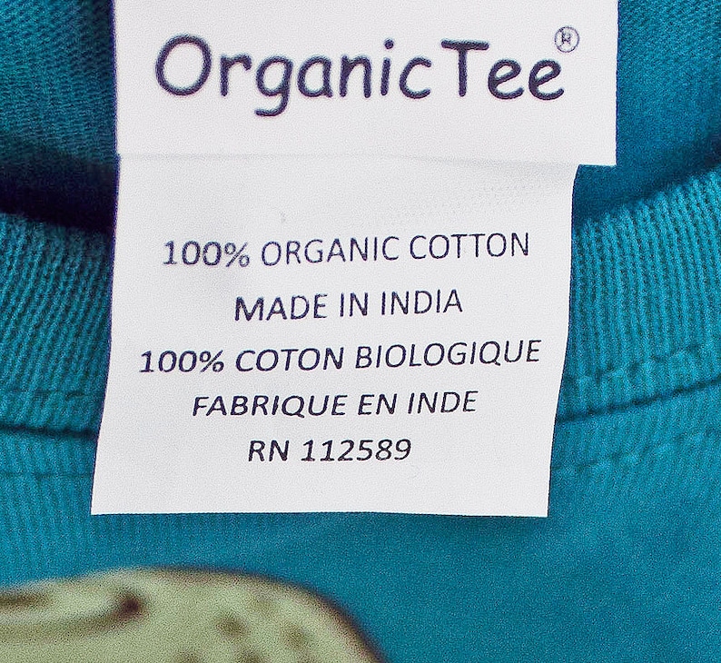 Może przedstawiać: Biała metka z czarnym tekstem na turkusowej koszulce. Metka zawiera napis "Organic Tee", "100% Organic Cotton", "Made in India", "100% Coton Biologique", "Fabrique en Inde" i "RN 112589".