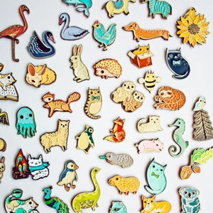May include: A collection of enamel pins featuring various animals, objects, and characters. The pins are colorful and detailed, with a variety of styles and designs. The pins are arranged in a grid pattern on a white background. The text "enamel pins" and "boygirlparty.com" are visible in the image.