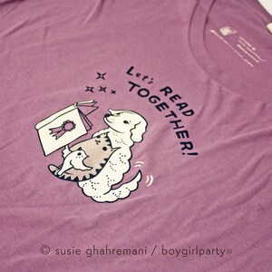 May include: Purple t-shirt featuring a white dog and cat reading a book. The text on the shirt reads "Let's Read Together!"