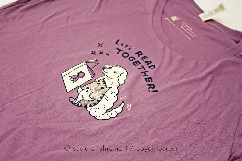 May include: Purple t-shirt featuring a white dog and cat reading a book. The text on the shirt reads "Let's Read Together!"