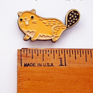May include: A yellow enamel pin of a cartoon beaver with a black tail. The beaver is about 1 inch long and is sitting on a wooden ruler with the words "Made in U.S.A." printed on it.