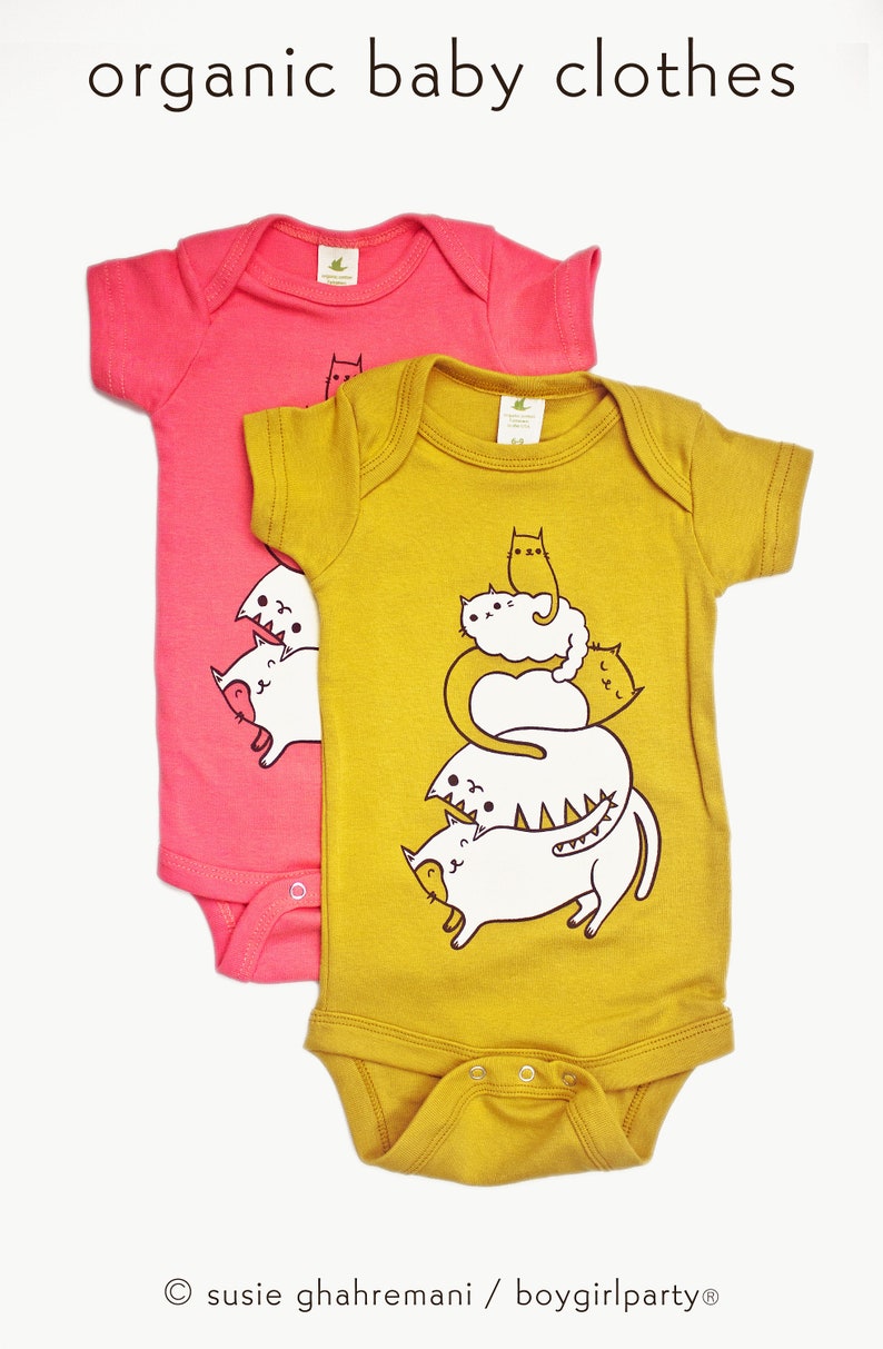 May include: Two organic cotton baby onesies. One is pink with a simple cat design. The other is mustard yellow with a stack of five cats, each with a different expression.