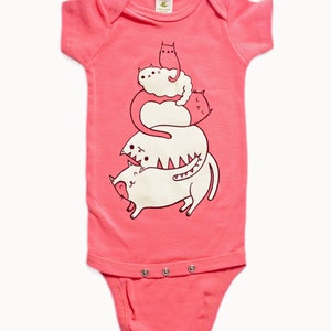 May include: Pink organic cotton baby onesie with a graphic of a stack of four cats. The cats are white with black outlines and are stacked on top of each other. The text "susie ghahremani / boygirlparty&reg;" is at the bottom of the onesie.