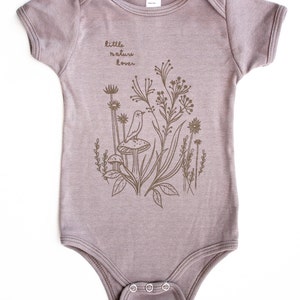 Puede incluir: Un body de beb&eacute; de color morado claro con un gr&aacute;fico marr&oacute;n de un p&aacute;jaro sentado en un hongo en un campo de flores. El texto "little nature lover" est&aacute; impreso en el body.