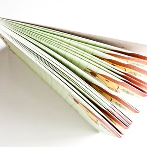Op de afbeelding: Een groen en bruin notitieboek met een bedrukte kaft. Het notitieboek is gesloten en waaiervormig opengevouwen, waardoor de pagina's zichtbaar zijn.