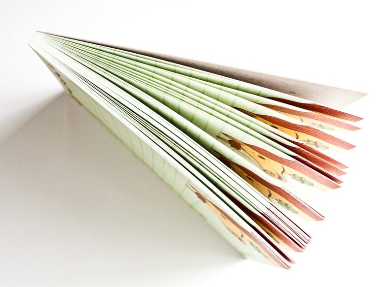 Op de afbeelding: Een groen en bruin notitieboek met een bedrukte kaft. Het notitieboek is gesloten en waaiervormig opengevouwen, waardoor de pagina's zichtbaar zijn.