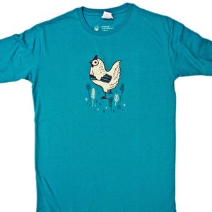 Może przedstawiać: Turkusowa organiczna koszulka z biało-czarną ilustracją kury na łące. Tekst "organic t-shirt" jest nadrukowany czarnym kolorem u g&oacute;ry obrazu.