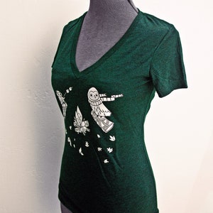 May include: A dark green v-neck t-shirt with a white graphic of owls flying with a scarf and leaves falling. The text "susie ghahremani / boygirlparty&reg;" is at the top of the image.