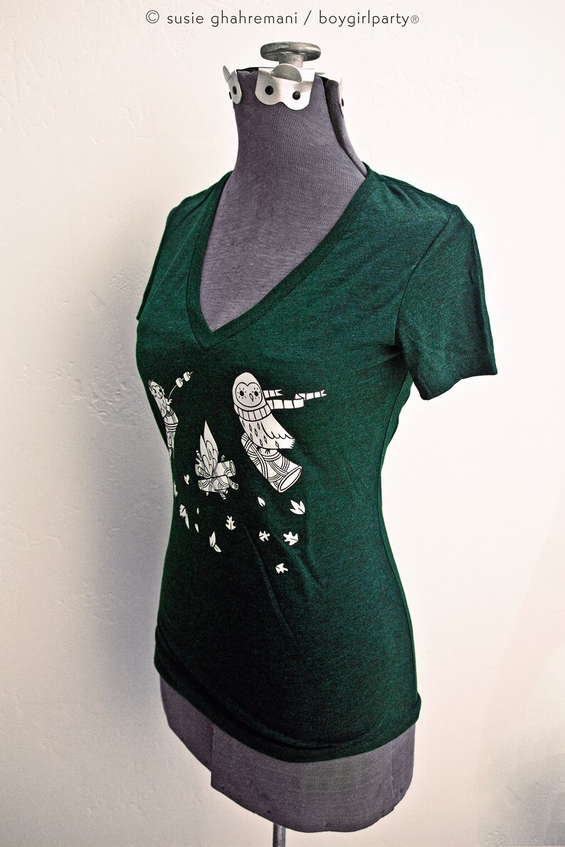 May include: A dark green v-neck t-shirt with a white graphic of owls flying with a scarf and leaves falling. The text "susie ghahremani / boygirlparty&reg;" is at the top of the image.
