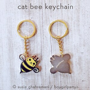 May include: A gold keychain with a bee-shaped charm. The bee is yellow and black with a cat face. The back of the charm is plain gold. The keychain is labeled "cat bee keychain" and "susie ghahremani/boygirlparty".