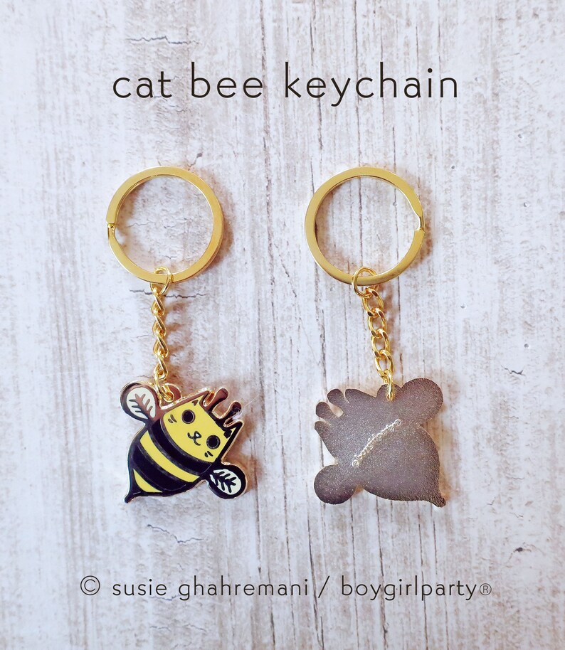 May include: A gold keychain with a bee-shaped charm. The bee is yellow and black with a cat face. The back of the charm is plain gold. The keychain is labeled "cat bee keychain" and "susie ghahremani/boygirlparty".