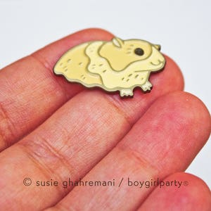 May include: A gold-colored enamel pin featuring a cartoon hamster with a light yellow body and a dark brown eye. The hamster is facing right and has a slightly surprised expression.