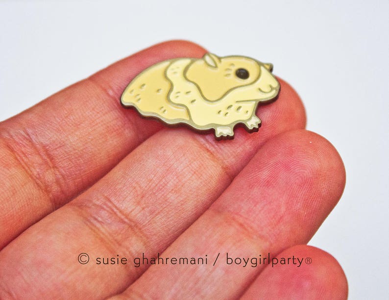 May include: A gold-colored enamel pin featuring a cartoon hamster with a light yellow body and a dark brown eye. The hamster is facing right and has a slightly surprised expression.