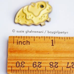 May include: A yellow enamel pin of a hamster, approximately 1 inch in size, with a black eye and a white background.
