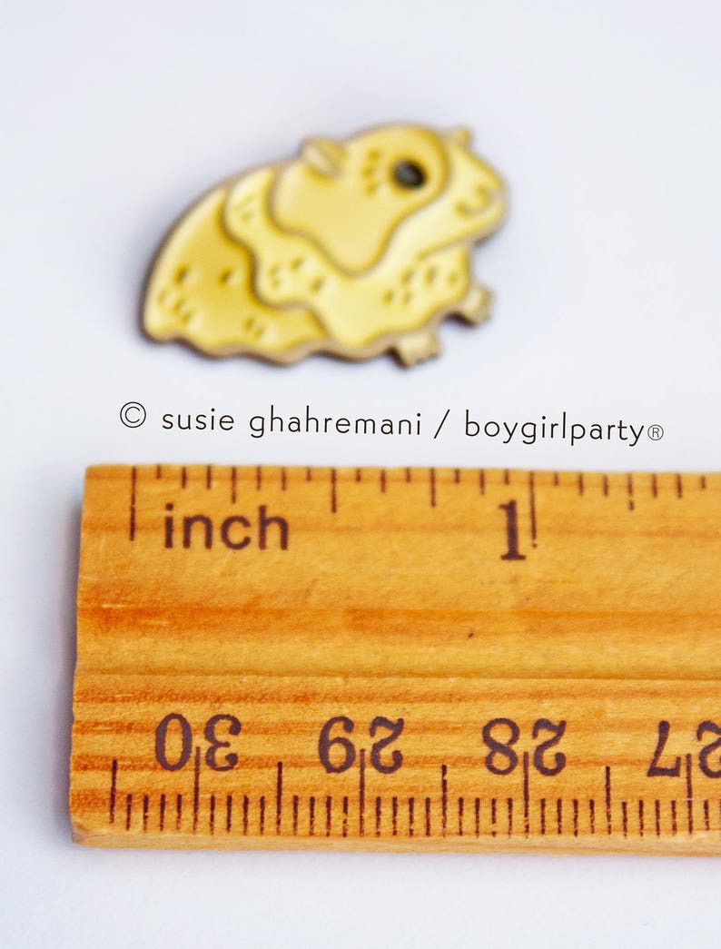 May include: A yellow enamel pin of a hamster, approximately 1 inch in size, with a black eye and a white background.