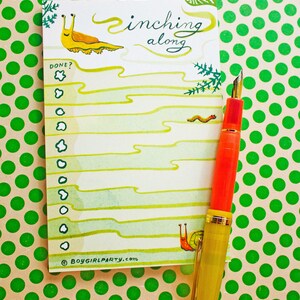 May include: A white to-do list notepad with a green and yellow snail illustration and the text "inching along". The notepad has a checklist with the word "DONE?" at the top. A gold and red fountain pen is lying on top of the notepad. The notepad is on a green polka dot background.
