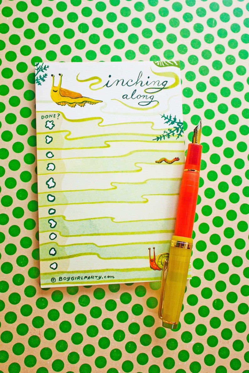 May include: A white to-do list notepad with a green and yellow snail illustration and the text "inching along". The notepad has a checklist with the word "DONE?" at the top. A gold and red fountain pen is lying on top of the notepad. The notepad is on a green polka dot background.