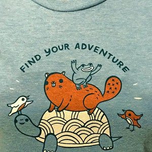 May include: A teal blue t-shirt with a graphic of a turtle, a beaver, a frog, and two birds. The text "Find Your Adventure" is printed in a semi-circle above the animals. The text "Suzie Ghahremani/BoyGirlParty for Artisans" is printed in the bottom right corner.