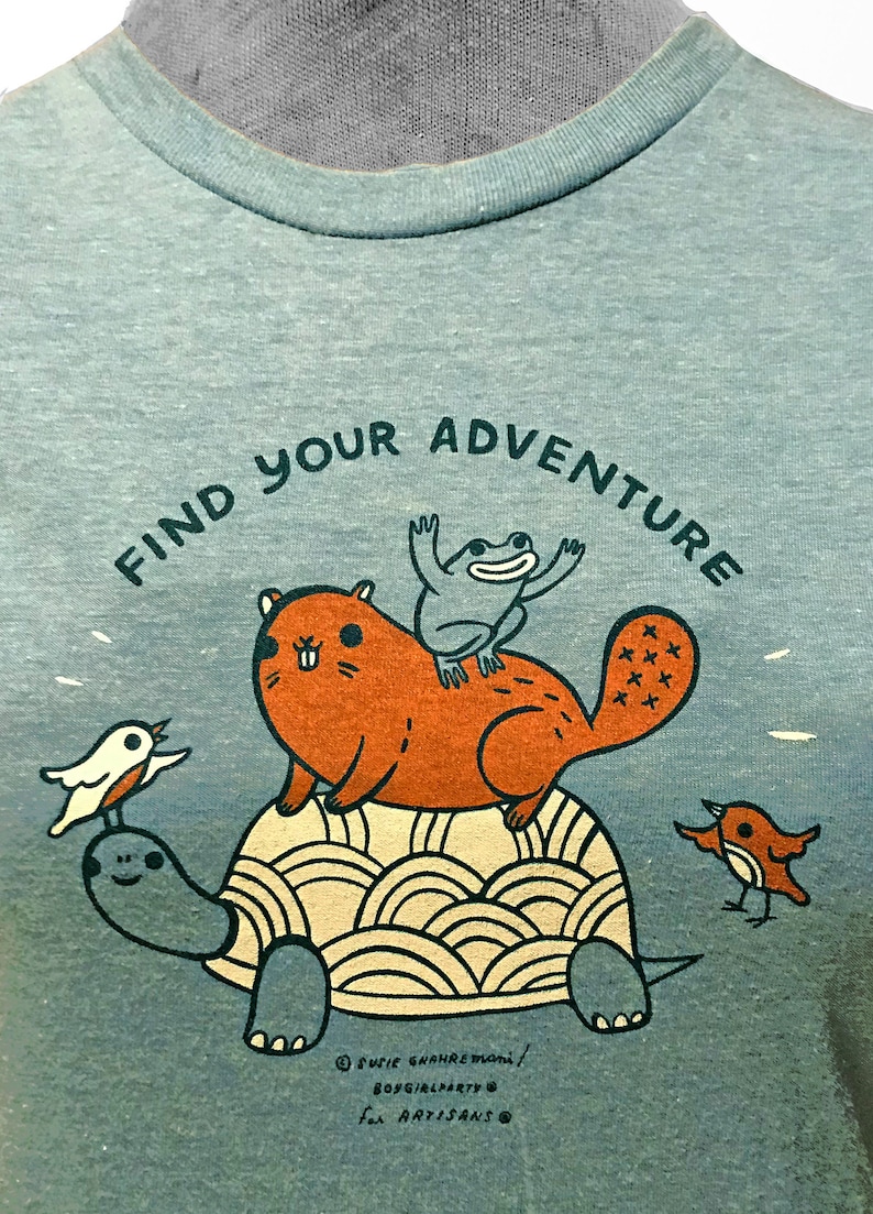 May include: A teal blue t-shirt with a graphic of a turtle, a beaver, a frog, and two birds. The text "Find Your Adventure" is printed in a semi-circle above the animals. The text "Suzie Ghahremani/BoyGirlParty for Artisans" is printed in the bottom right corner.