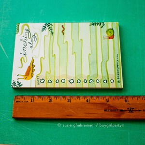 May include: A white notepad with a green and yellow illustration of a snail inching along. The notepad has a checklist with the words "Inchling along" and "Done?" written on it. The notepad is 6 inches long.