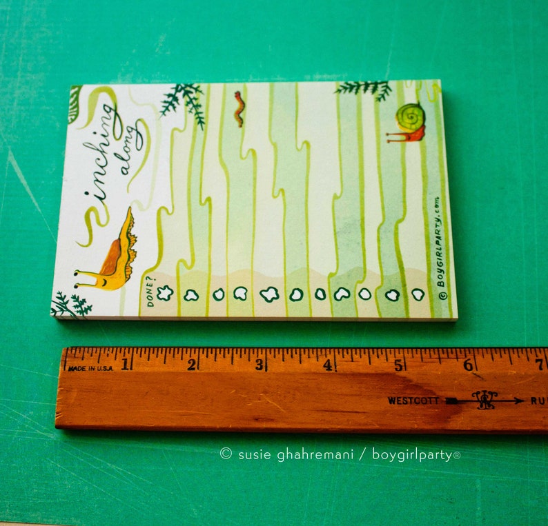 May include: A white notepad with a green and yellow illustration of a snail inching along. The notepad has a checklist with the words "Inchling along" and "Done?" written on it. The notepad is 6 inches long.